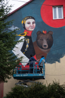 Dwie osoby tworzą mural. Kobieta w stroju ludowym, za nią niedźwiedź na ścianie budynku.