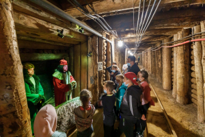 Grupa dzieci w maseczkach stoją w kopalni  i patrzą się na figurki w przebraniach z naklejonymi twarzami.