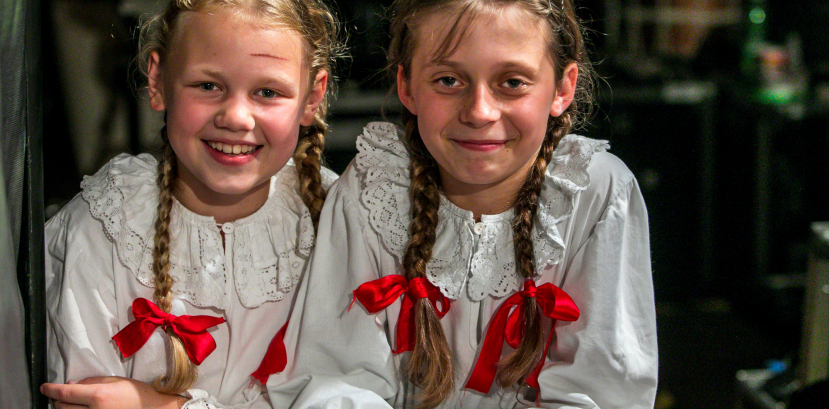 dwie dziewczynki w strojach regionalnych uśmiechają się i opierają o scenę