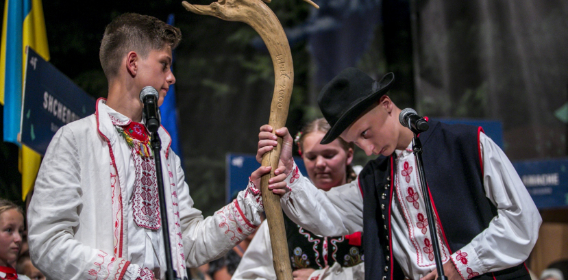 Dwóch chłopców w strojach ludowych na scenie, przy mikrofonie. Trzymają drewniany kij.