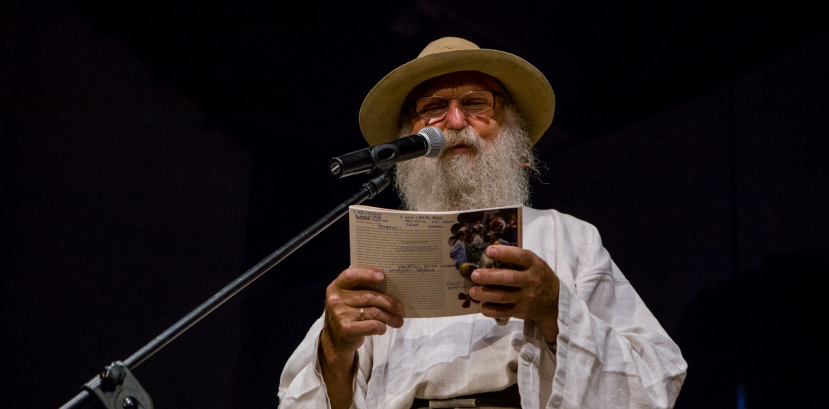 Mężczyzna z brodą w kapeluszu stoi przy mikrofonie z otwartą książką w rękach.