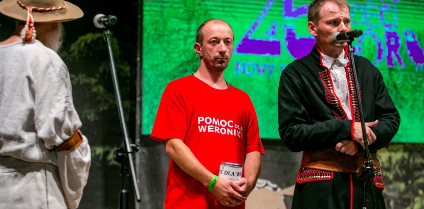 Trzech mężczyzn na scenie przy mikrofonach. Jeden z nich trzyma puszkę, a drugi ma kapelusz na głowie.