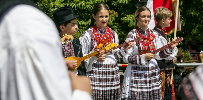 Dwie dziewczyny w takich samych strojach regionalnych stoją obok siebie. Za nimi widać zieleń, a przed nimi innych ludzi.