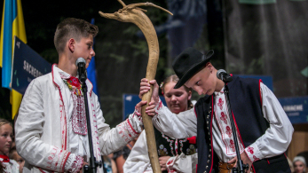 Dwóch chłopców w strojach ludowych na scenie, przy mikrofonie. Trzymają drewniany kij.