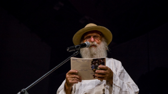 Mężczyzna z brodą w kapeluszu stoi przy mikrofonie z otwartą książką w rękach.