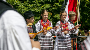 Dwie dziewczyny w takich samych strojach regionalnych stoją obok siebie. Za nimi widać zieleń, a przed nimi innych ludzi.