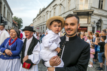 Ksiądz trzyma dziecko na rękach. Z tyłu idą ludzie.