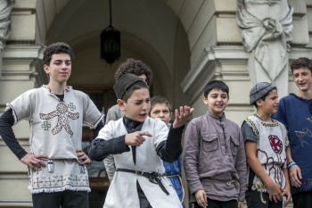 Chłopcy stoją w rzędzie na płycie rynku. Jeden z nich udaje, że trzyma broń i strzela.