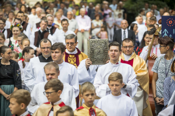 Księża i ministranci idą przez aleję między wypełnionymi ludźmi ławkami ustawionymi na placu przykościelnym