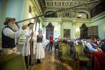 Na pierwszym planie starszy mężczyzna w regionalnym stroju stoi obok trombity. W tle sala wypełniona ludźmi