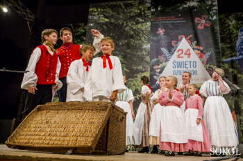 Grupa dzieci w strojach ludowych na scenie. Chłopcy stoją przy wiklinowej skrzyni.