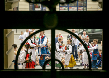 Za oknem grupa młodzieży w strojach ludowych tańczą ze sobą.