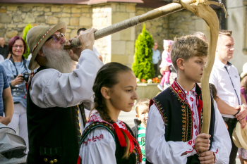 Mężczyzna gra na drewnianym instrumencie, a obok niego stoi dziewczyna i chłopak w strojach ludowych.