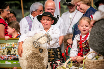 Chłopczyk i dziewczynka w strojach ludowych siedzą. Chłopczyk trzyma sztucznego baranka.